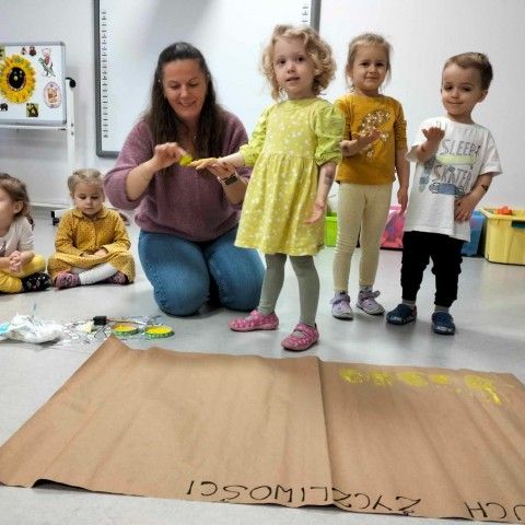 Powiększ obraz: Grupa dzieci stoi przed plakatem, na którym za chwilę odciśnięte zostaną kształty ich dłoni, tworząc łańcuszek życzliwości