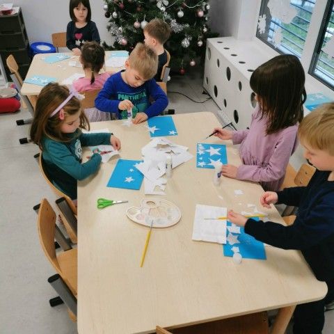 Powiększ obraz: Dzieci siedzą przy stolikach, wycinają oraz przyklejają papierowe gwiazdki 