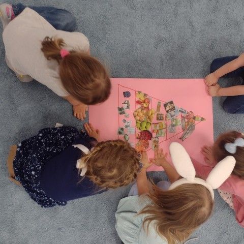 Powiększ obraz: Dzieci trzymają na dywanie plakat z piramidą żywienia
