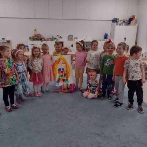 Powiększ obraz: Uśmiechnięte dzieci stoją w półokręgu i prezentują swoją wspólną pracę plastyczną – portret Pani Wiosny